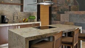 Cozinha com ilha central, cadeiras de madeira, forno embutido e painel de fundo de pedra.