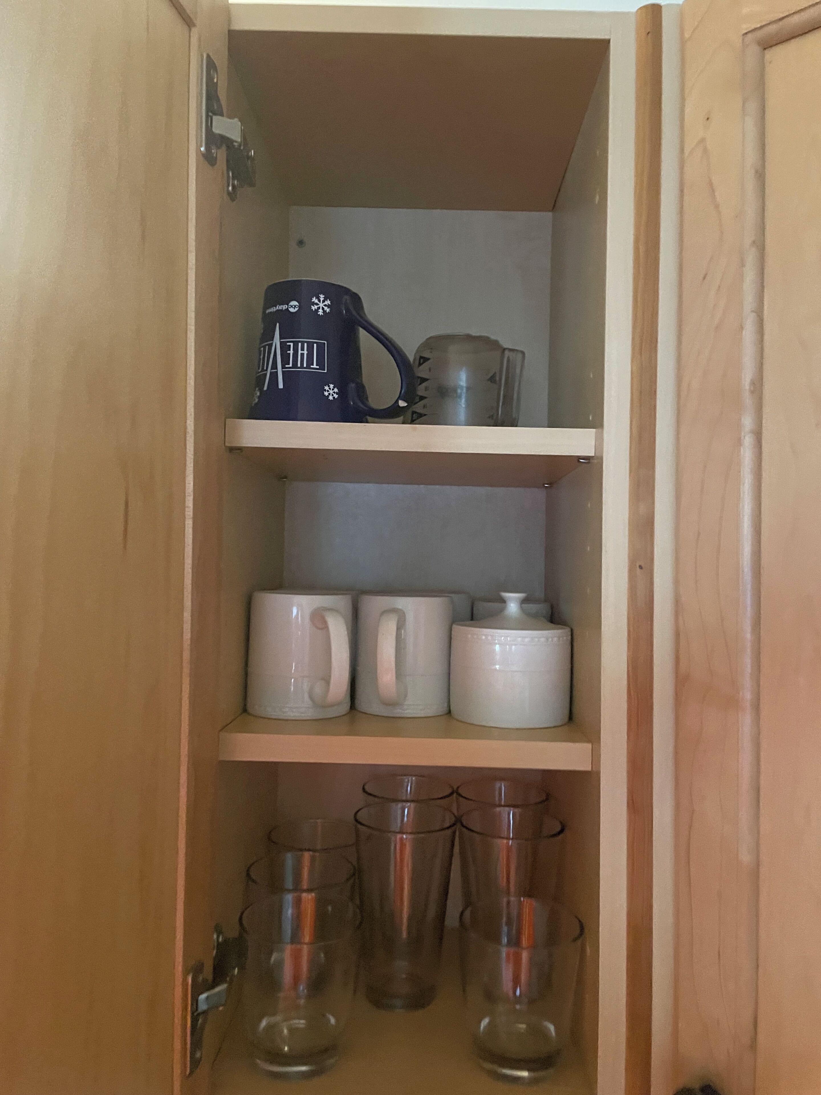 A wooden cabinet with shelves holding mugs, a glass container, and clear glasses.