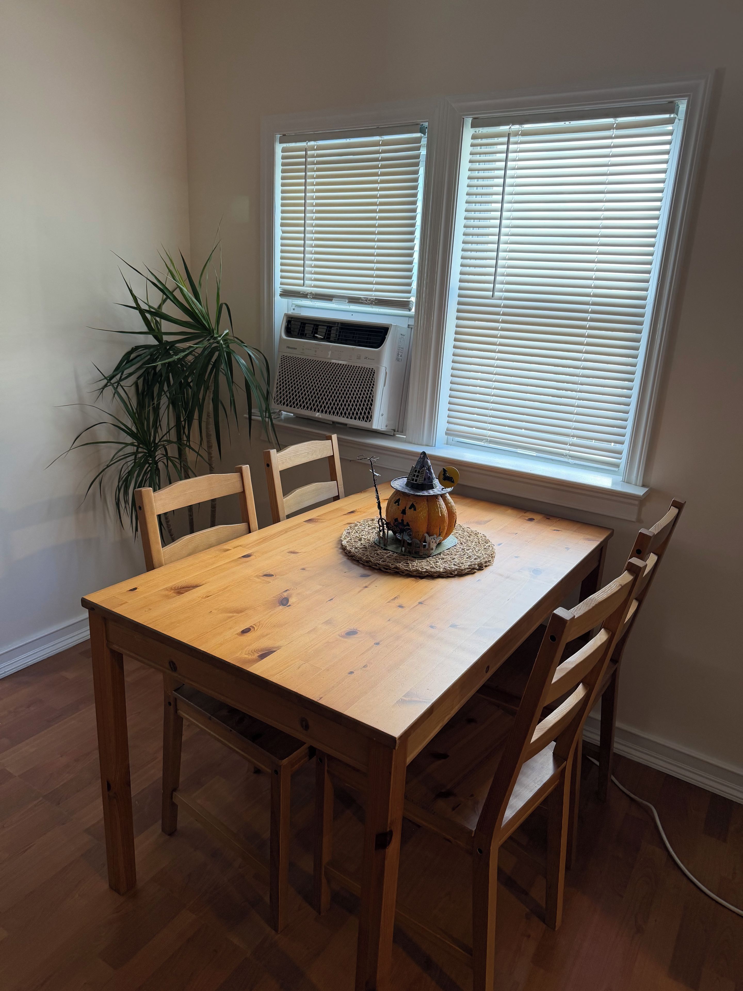 Un comedor con una mesa de madera, cuatro sillas, un centro decorativo y un ventanal con persianas.