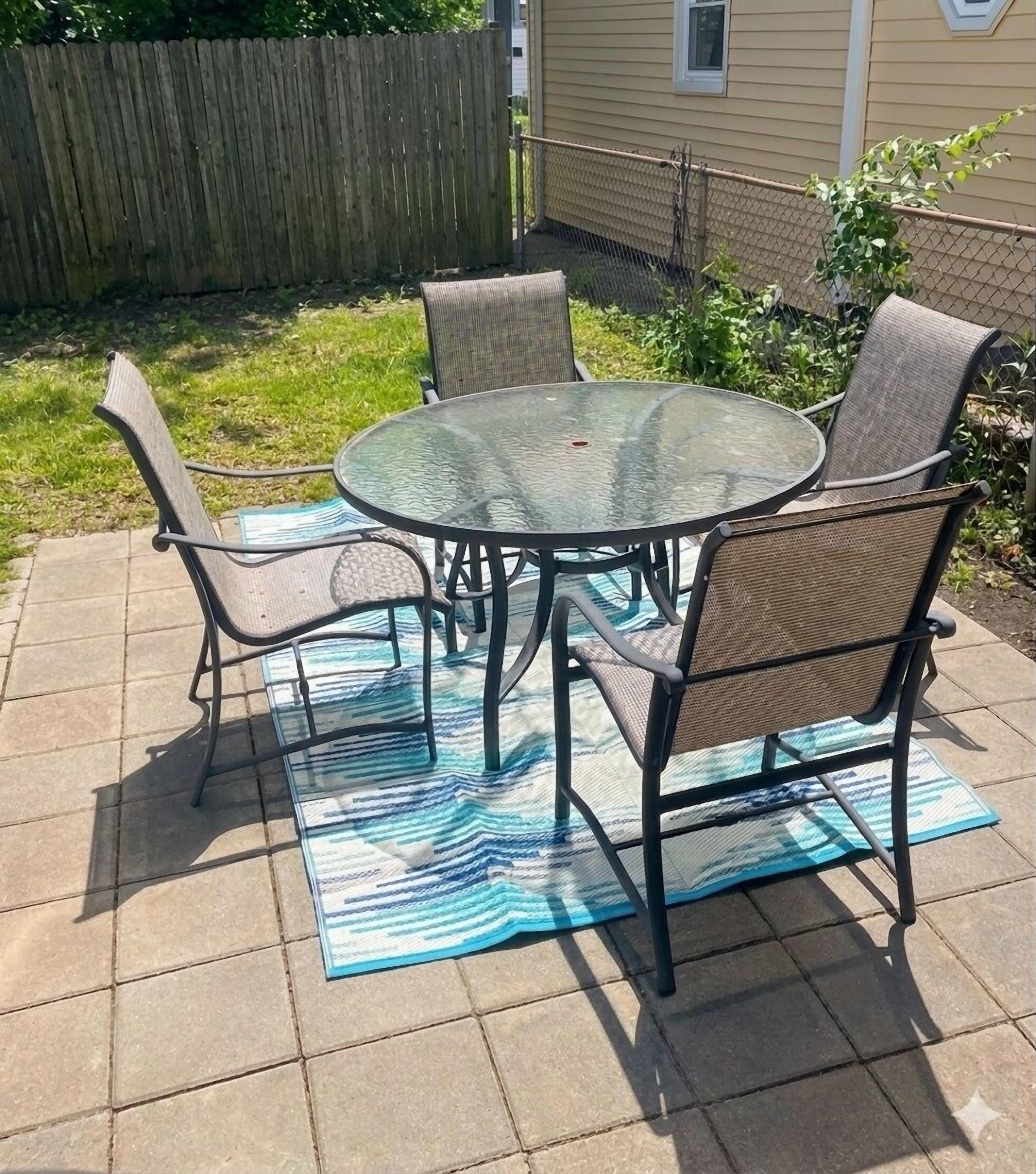 Un patio con una mesa de vidrio y cuatro sillas, rodeado por una cerca de madera y una casa.
