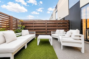 A patio area with a white sofa, white armchairs, and a small table, surrounded by artificial green grass and a wooden fence. - Salisbury Garden | 2BR with Parking | CBD (Christchurch)