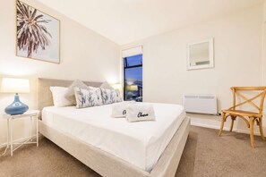 A hotel room with a bed, a nightstand with a lamp, a chair, a window with a view, and a wall-mounted air conditioner.