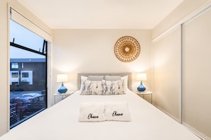 A bedroom with a bed, two bedside tables, a wall-mounted circular decoration, and a window with a view of buildings and parked cars. - Salisbury Garden | 2BR with Parking | CBD (Christchurch)