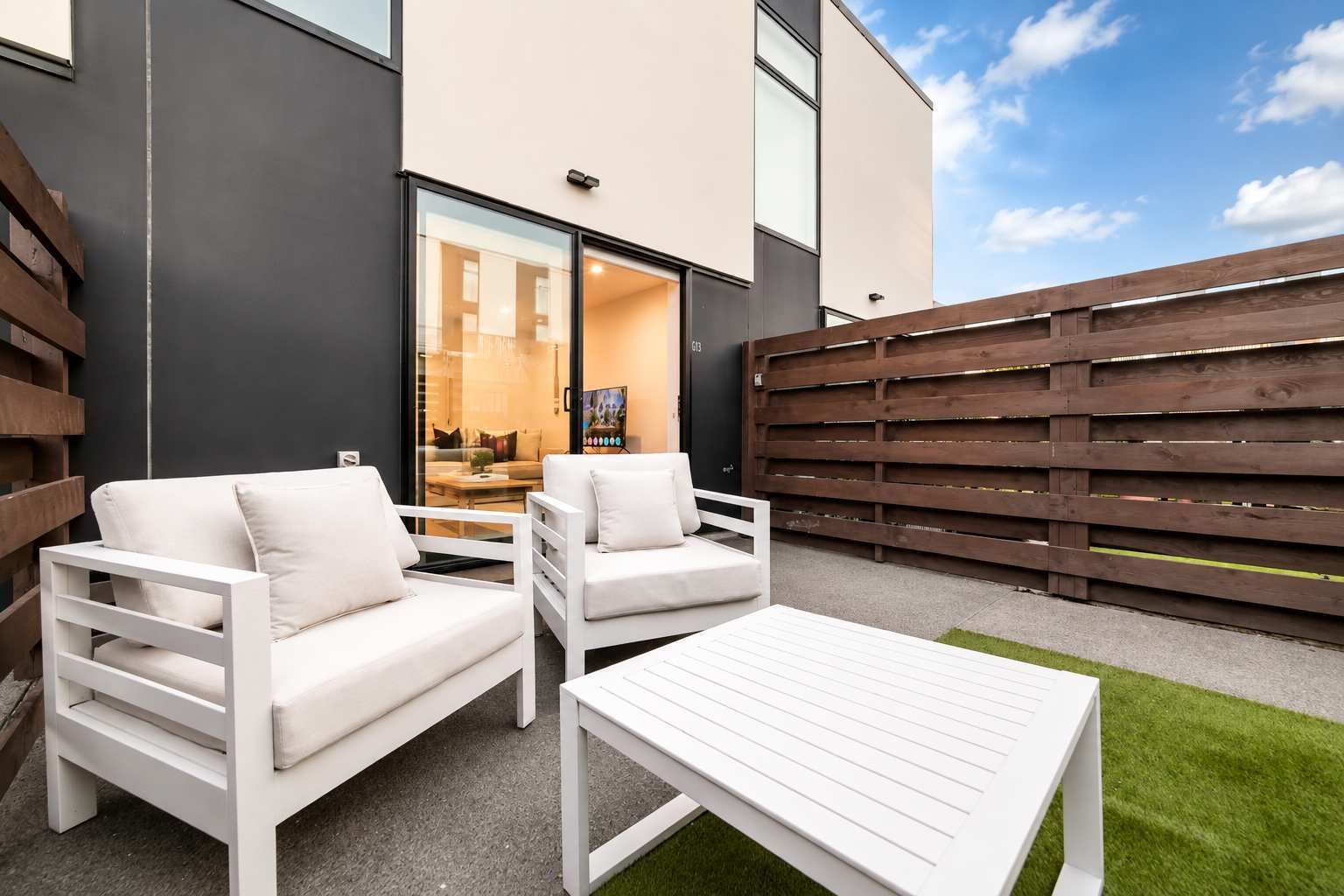 A modern outdoor seating area with two white sofas and a white table, set against a building with a dark facade and a wooden fence.