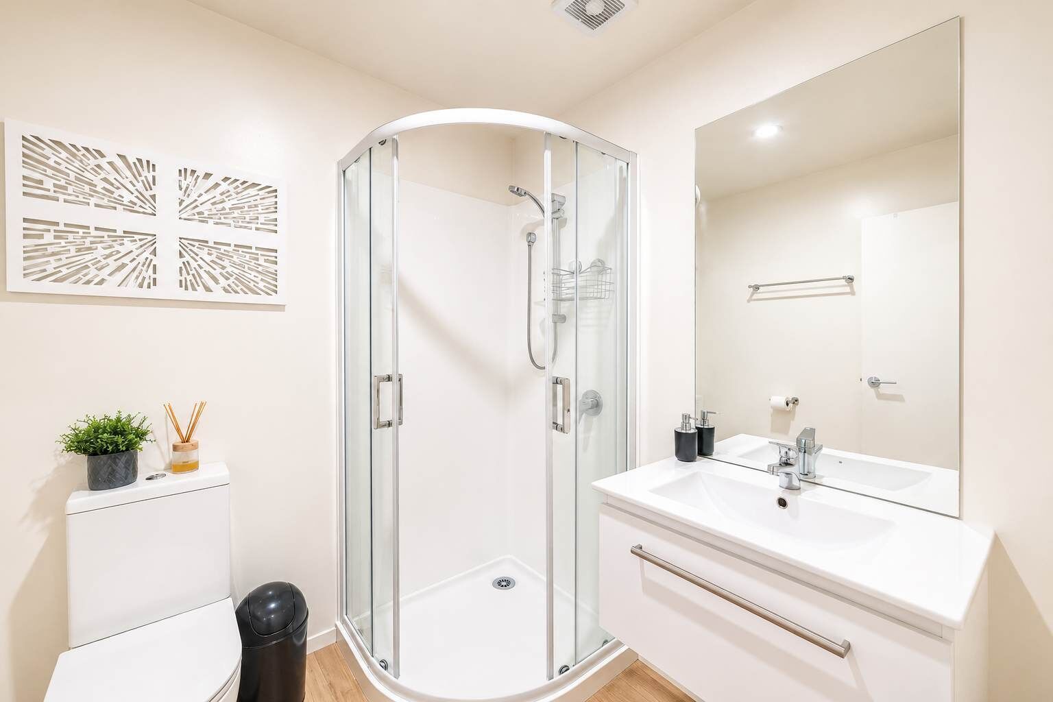 Un baño moderno con cabina de ducha de vidrio, un lavamanos blanco con un espejo grande y una pequeña planta en maceta sobre el mueble.