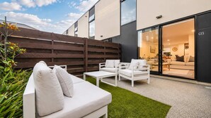 An outdoor seating area with a white sofa and chairs, a wooden fence, and a view into a modern interior space. - Salisbury Garden | 2BR with Parking | CBD (Christchurch)