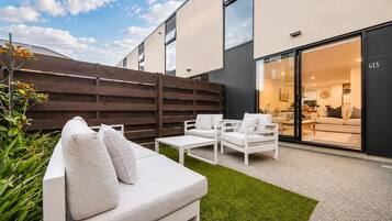 An outdoor seating area with a white sofa and chairs, a wooden fence, and a view into a modern interior space.