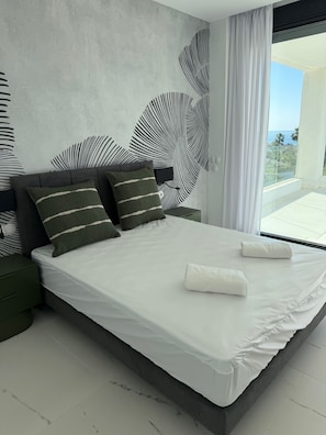 Ein modernes Schlafzimmer mit einem großen Bett, dunkelgrünen Nachttischen und Meerblick vom Balkon.