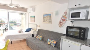 A room with a bed, a grey sofa, a small kitchenette with a microwave, a toaster, and a window with a city view. - Discover this charming studio located in Santa Margarita, Roses! (Roses)