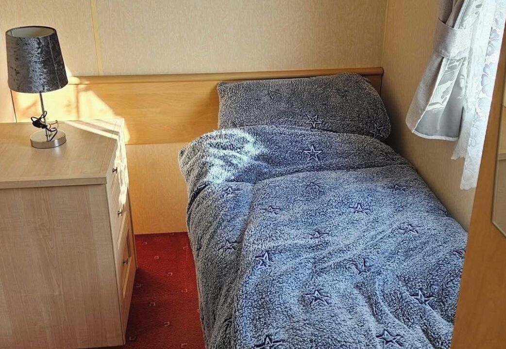 Una cama individual con una manta estampada con estrellas, una lámpara de noche, una mesita de noche de madera y un suelo cubierto de alfombra roja.