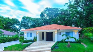 Una casa a un piano con tetto in tegole rosse, un ingresso principale con gradini e un prato curato con piccole palme.