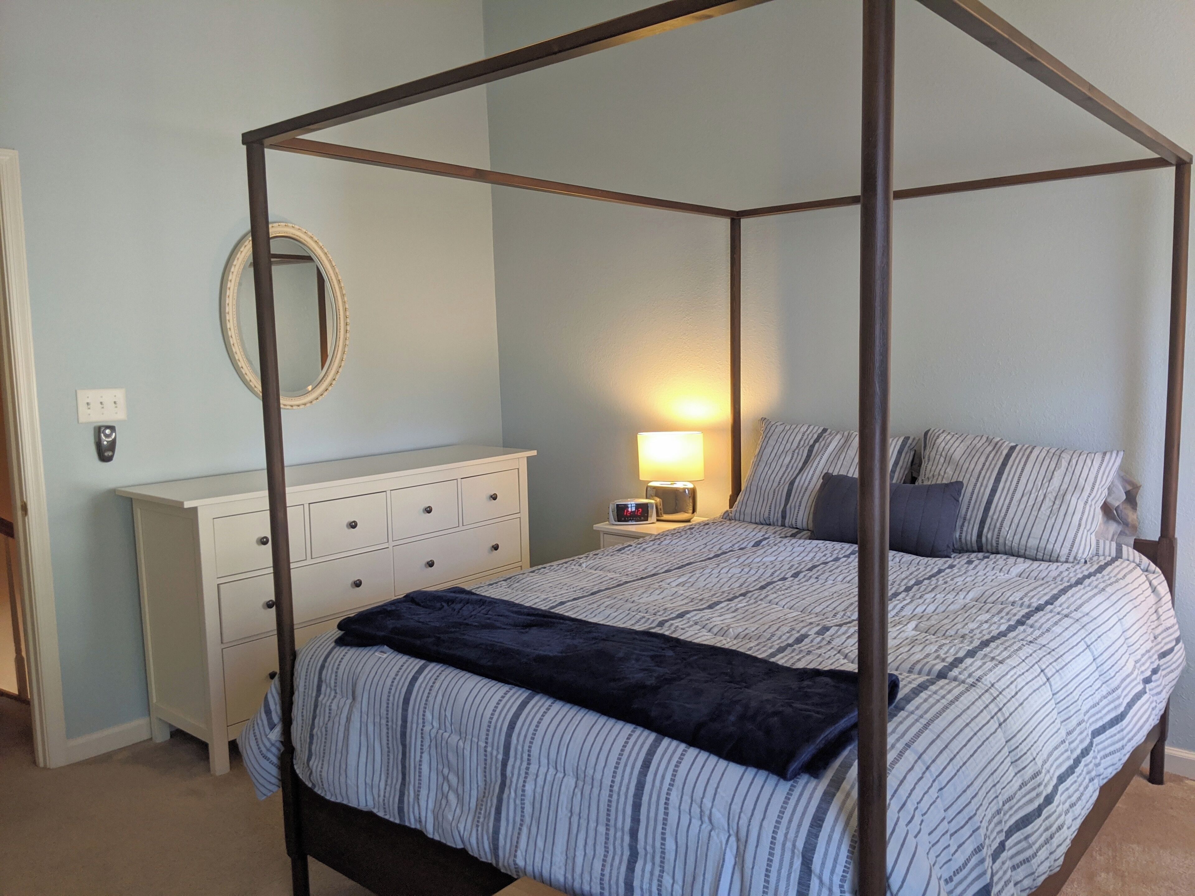 A four-poster bed with a canopy, a bedside table with a lamp, a dresser, and a mirror on the wall.