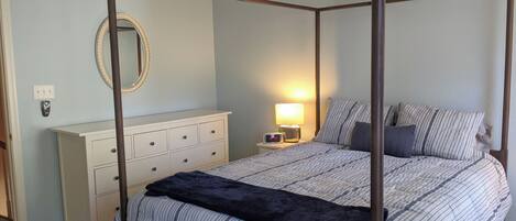 A four-poster bed with a canopy, a bedside table with a lamp, a dresser, and a mirror on the wall.