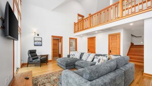 Ein geräumiger Wohnbereich mit grauem Ecksofa, Holzboden, einer Holztreppe und einem an der Wand montierten Fernseher.