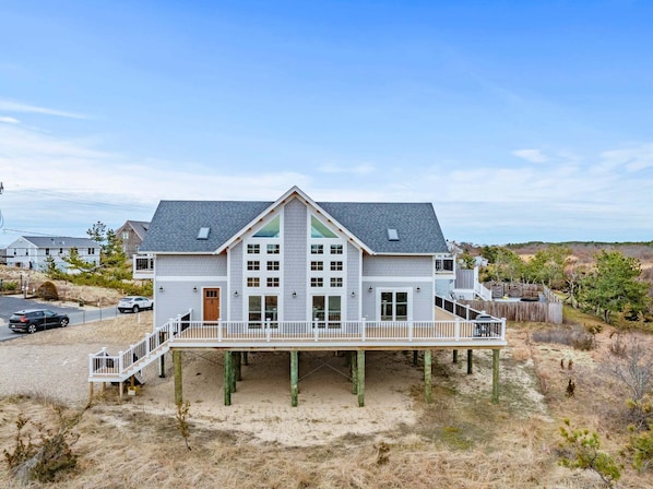 A two-story house with a balcony, wooden deck, and a sandy beach setting.