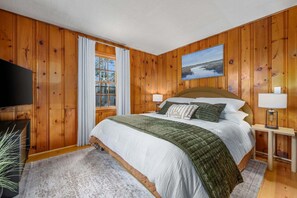 A wooden-paneled bedroom with a bed, two bedside lamps, a television, and a window with curtains.