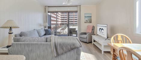 Un salon comprenant un canapé, une table à manger, un ventilateur de plafond et une vue sur un immeuble, visible à travers une porte vitrée.