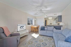 Ein Wohnzimmer mit einer Couch, einem Esstisch mit Stühlen, einem Deckenventilator, einer Küchenzeile mit Schränken und einem Schild, das Urlaubswohnungen bewirbt.
