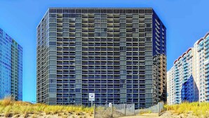 A high-rise building with a grid of windows, surrounded by other buildings and a sandy area with grass. - Golden Sands 1106: Gorgeous Oceanfront 1BR condo | Fully renovated and decorated (Ocean City)