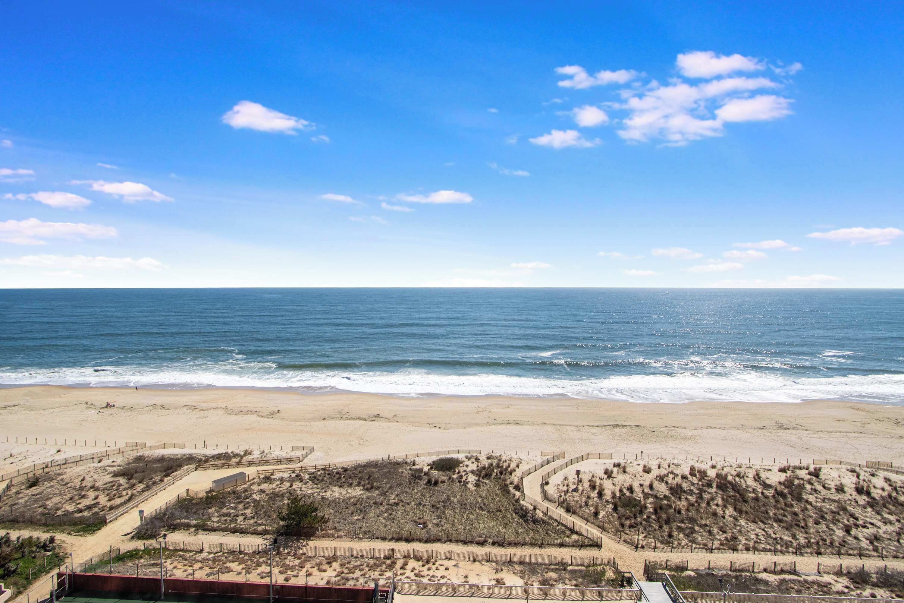 Une plage avec un ciel dégagé, un rivage sablonneux et une zone délimitée par une clôture, où l’on trouve de la végétation.