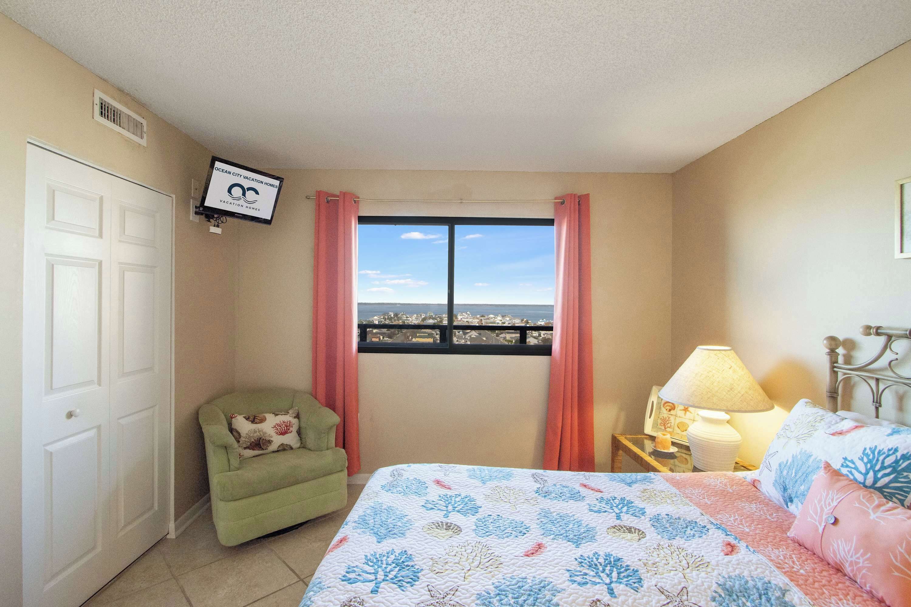A bedroom with a bed, a chair, a nightstand with a lamp, a window with curtains, and a TV mounted on the wall.