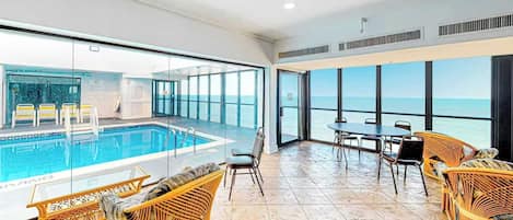 A pool area with a view of the ocean, wicker furniture, and a table with chairs.