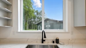 Eine Küche mit Spüle aus Edelstahl, einem schwarzen Wasserhahn und einem Fenster mit Blick auf ein Haus und Bäume.