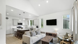 Eine moderne Küche mit weißen Schränken, einer zentralen Kochinsel und Barhocker. Es gibt einen Wohnbereich mit einer Couch, einem Couchtisch und einem an der Wand befestigten Fernseher.