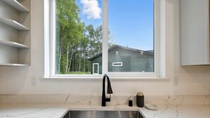 Eine Küche mit Spüle aus Edelstahl, einem schwarzen Wasserhahn und einem Fenster mit Blick auf ein Haus und Bäume.