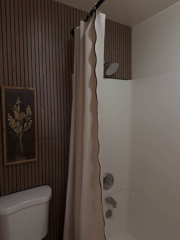A bathroom with a wooden panel wall, a white toilet, a shower with a white curtain, and a framed floral artwork.