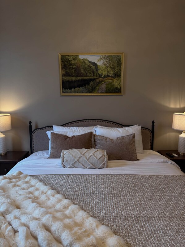 Una cama bien hecha con un edredón estampado, un cabecero con detalles decorativos, un cuadro de paisaje enmarcado sobre la cama y dos lámparas de noche a cada lado.