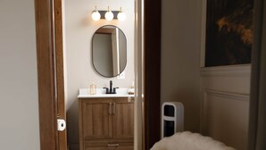 A room with a wooden door leading to a bathroom with a mirror, sink, and vanity. There is a white chair with a fluffy blanket in the foreground.