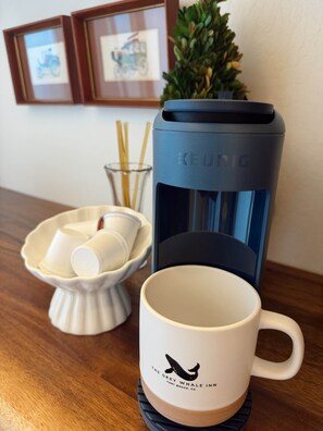 A coffee maker, a mug with a whale logo, and a bowl of cups on a wooden table. - The Noyo Ridge Suite (Fort Bragg)