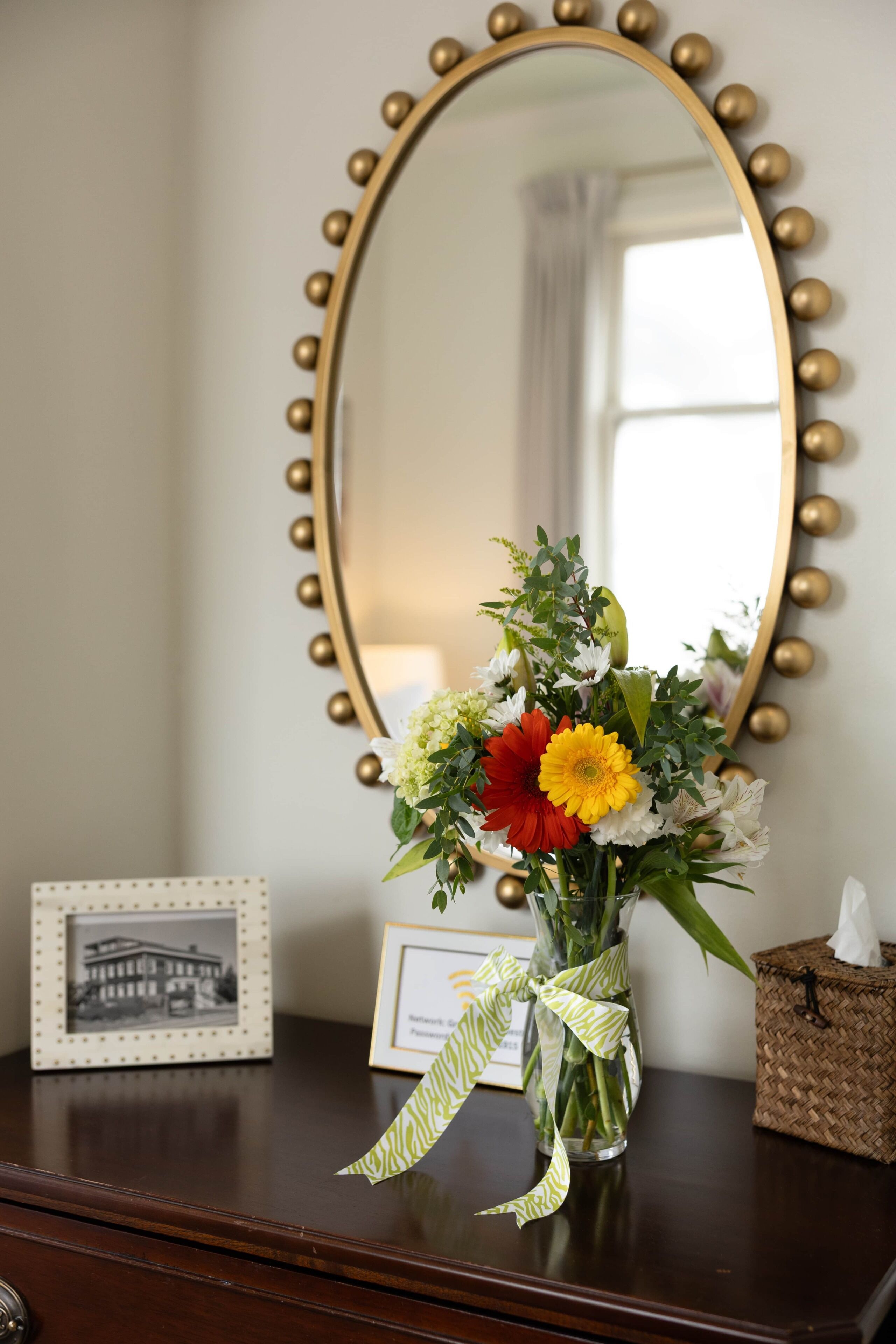 Et spejl med gylden ramme og dekorative kugler, en vase med blomster, et billedramme og en tissueske på en kommode.