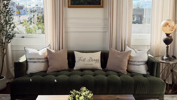Ein grünes Sofa mit einem Kissen mit dem Aufdruck 'Fort Bragg California', einem gerahmten Gemälde und einem Tisch mit Kerze und Blumenvase.