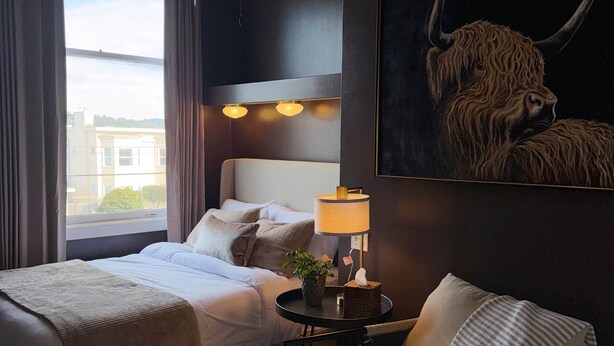 Ett hotellrum med en säng, en stol, en lampa, en tavla och utsikt över omgivningen.
