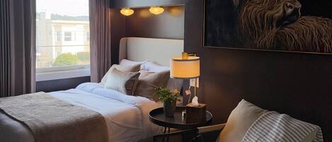 A hotel room with a bed, a chair, a lamp, a painting, and a view of the outside.