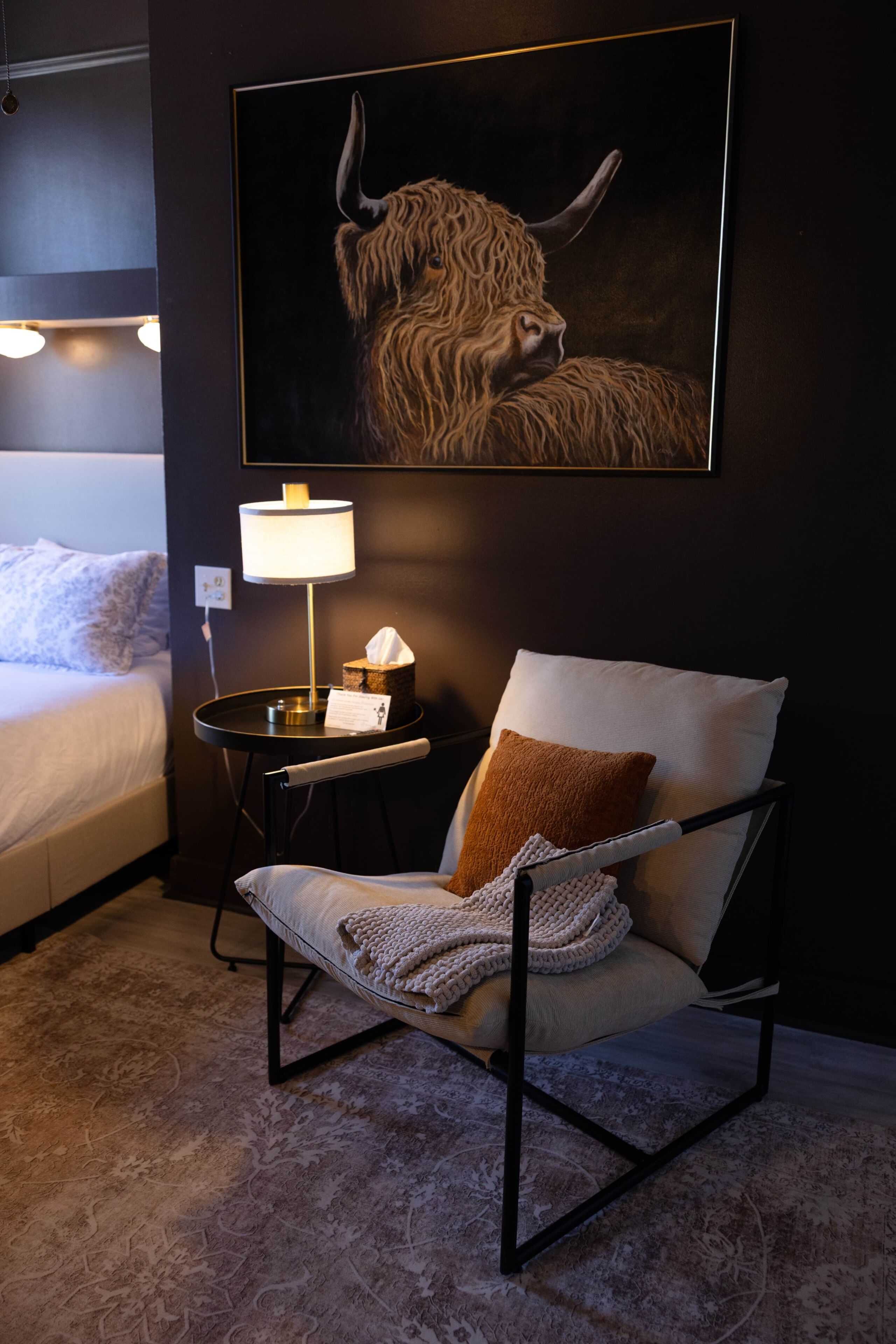 Hotellihuone, jossa on sänky, nojatuoli, jossa on päällä heittopeitto, yöpöytä lampun kanssa ja seinällä oleva härkän kuva.