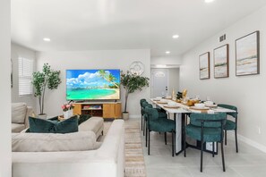 Una sala moderna con televisor de pantalla plana, un comedor con mesa blanca y sillas verdes, y un pasillo con puerta.