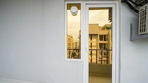 Eine weiße Außenwand mit einer Tür, einem Fenster und einem Balkon mit Metallgeländer, sowie Blick auf benachbarte Gebäude.