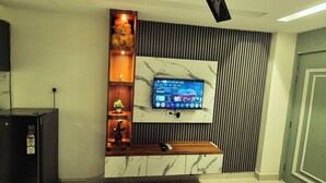 Ein modernes Wohnzimmer mit an der Wand montiertem Fernseher, einer eingebauten Holzregal mit Dekoration, einem schwarzen Kühlschrank und einer Tür rechts.