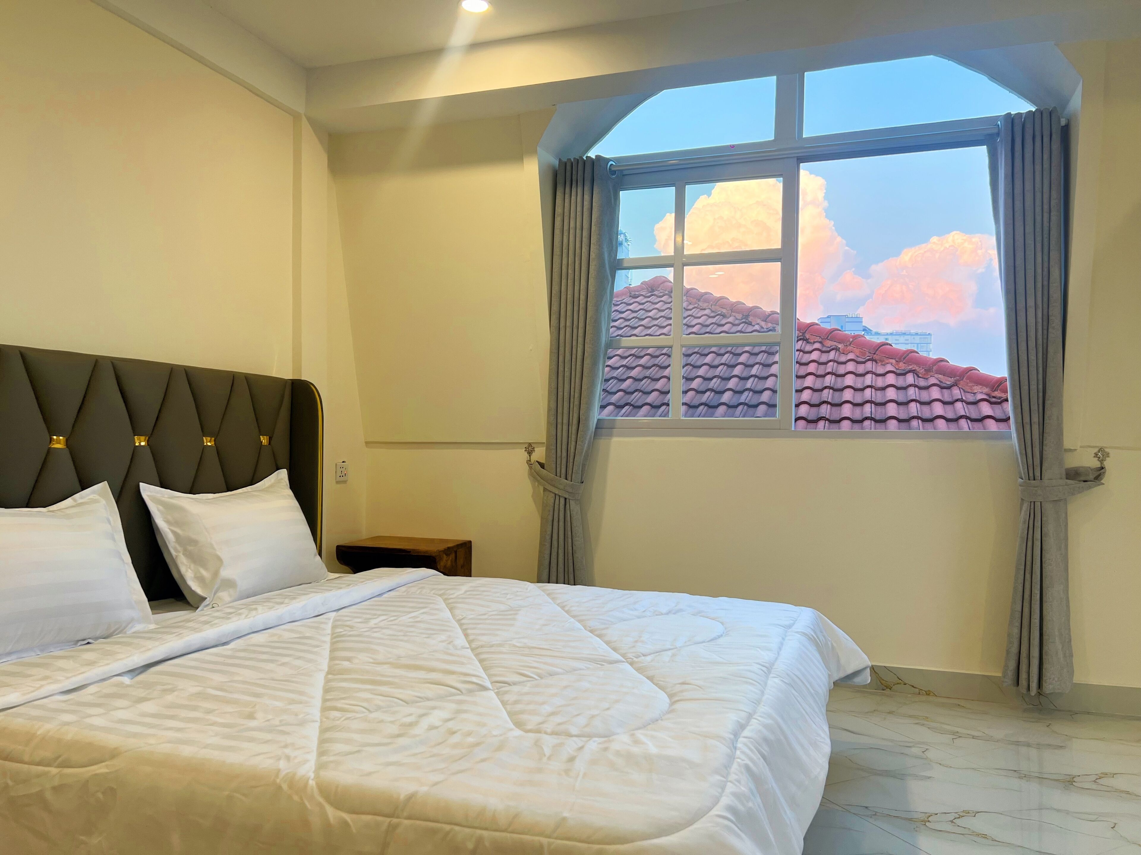 A hotel room with a bed, a nightstand, a window with curtains, and a view of a red-tiled roof.