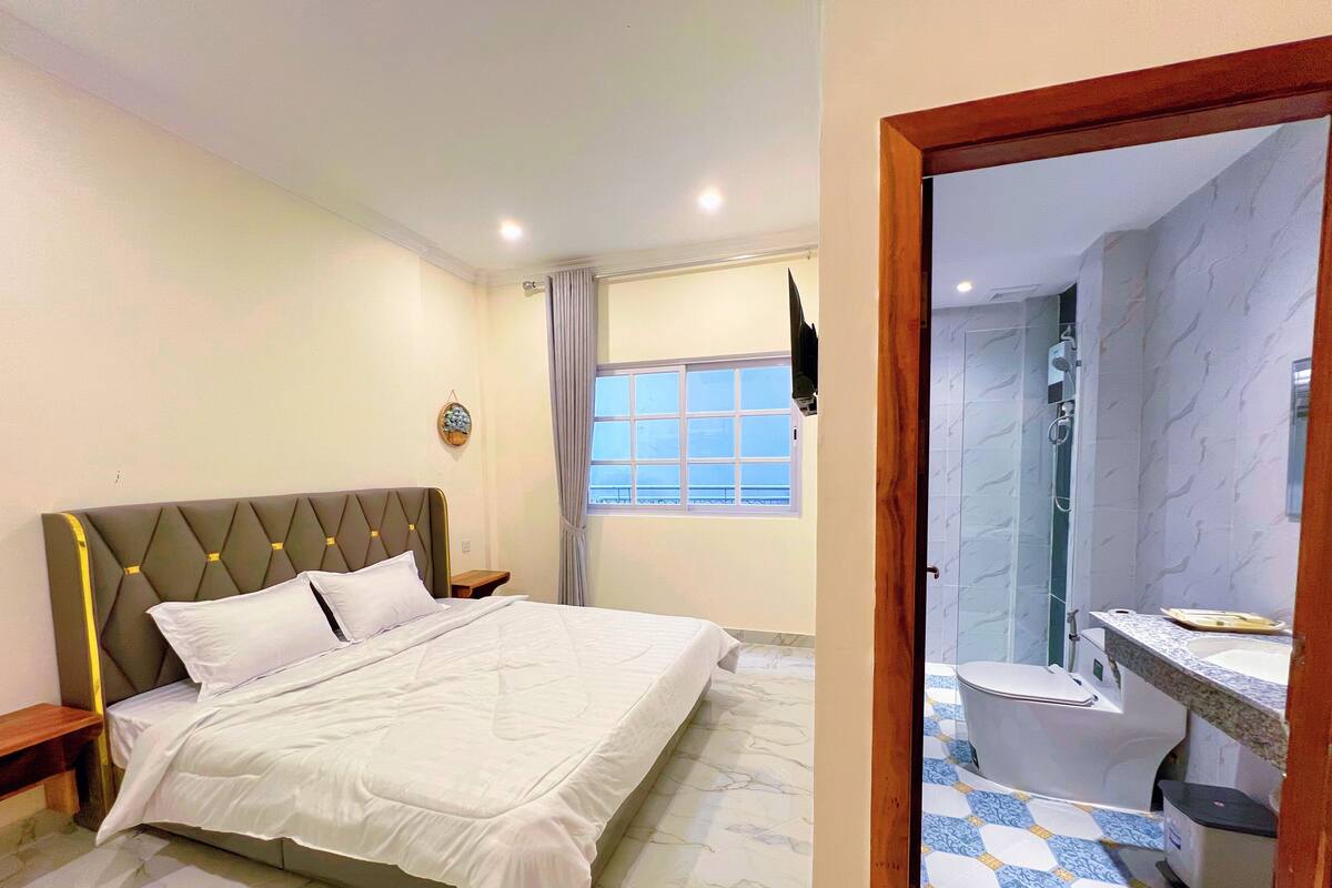 Un dormitorio con cama, mesita de noche, ventana con cortinas y baño completo con inodoro, lavabo y ducha.
