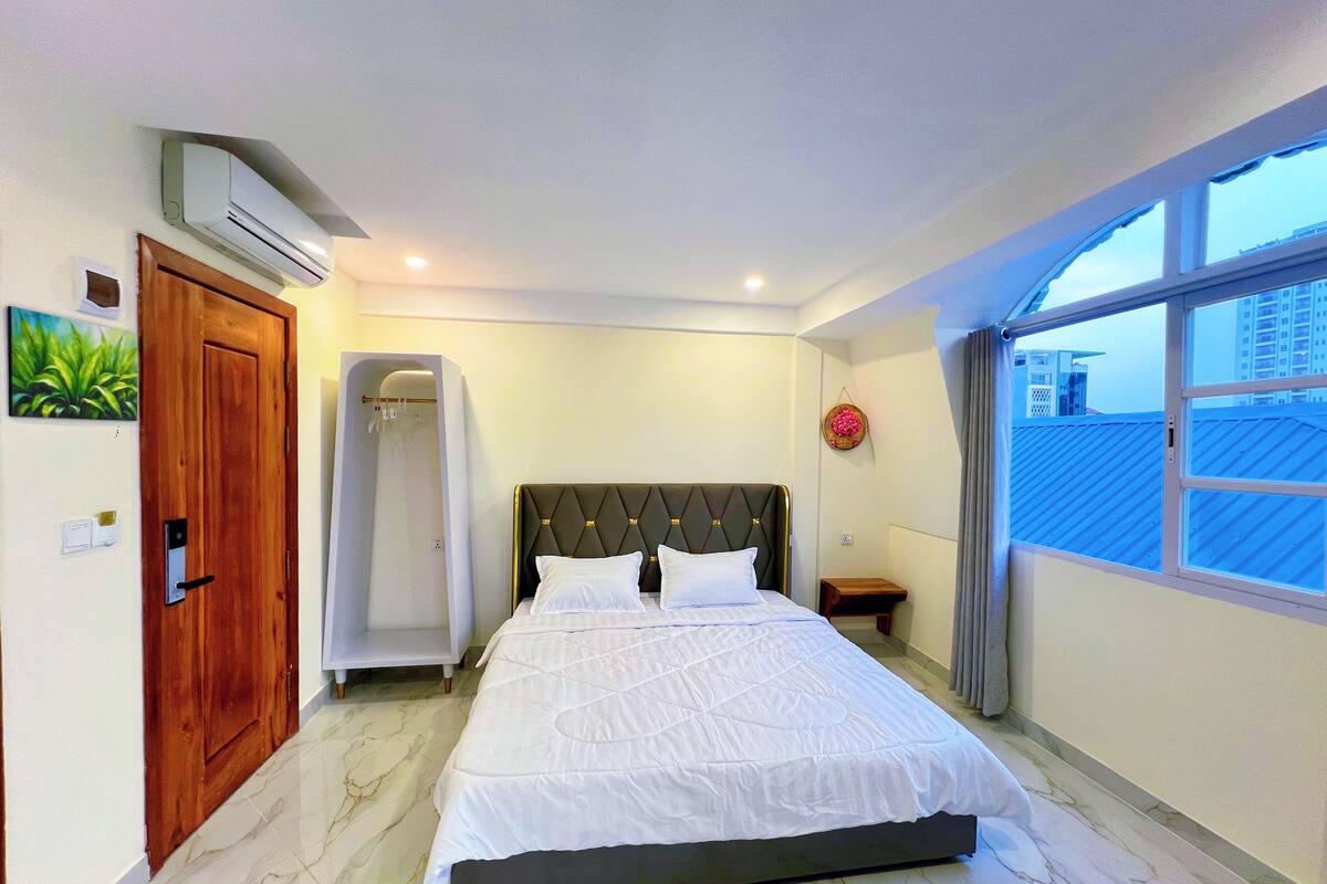 Un dormitorio con una cama, una puerta de madera, un equipo de aire acondicionado, una mesita pequeña y una ventana.