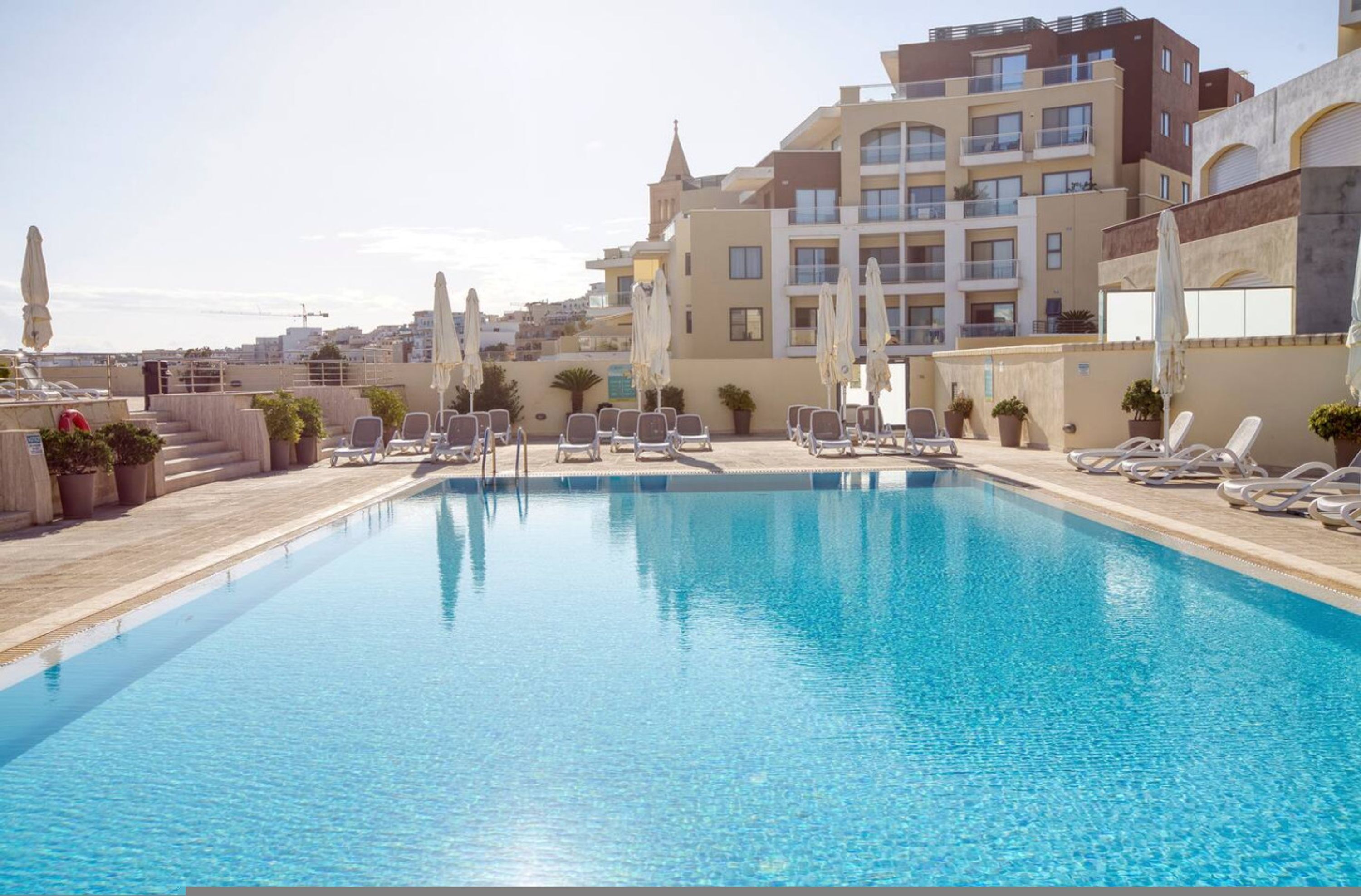 Une piscine entourée de bâtiments, avec des chaises longues et des parasols.