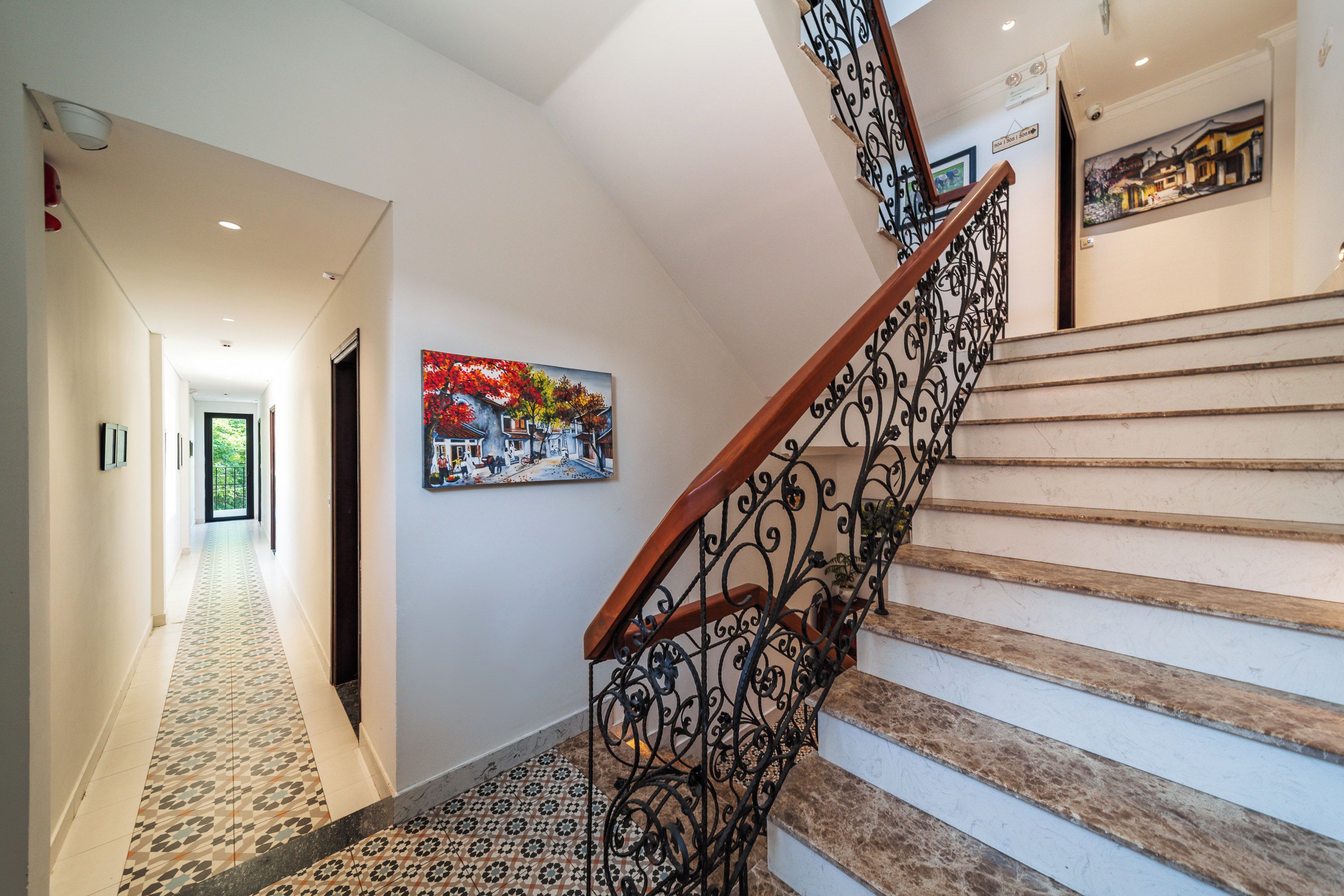 A staircase with a wooden handrail and black wrought iron balusters, leading to a landing with a colorful painting.