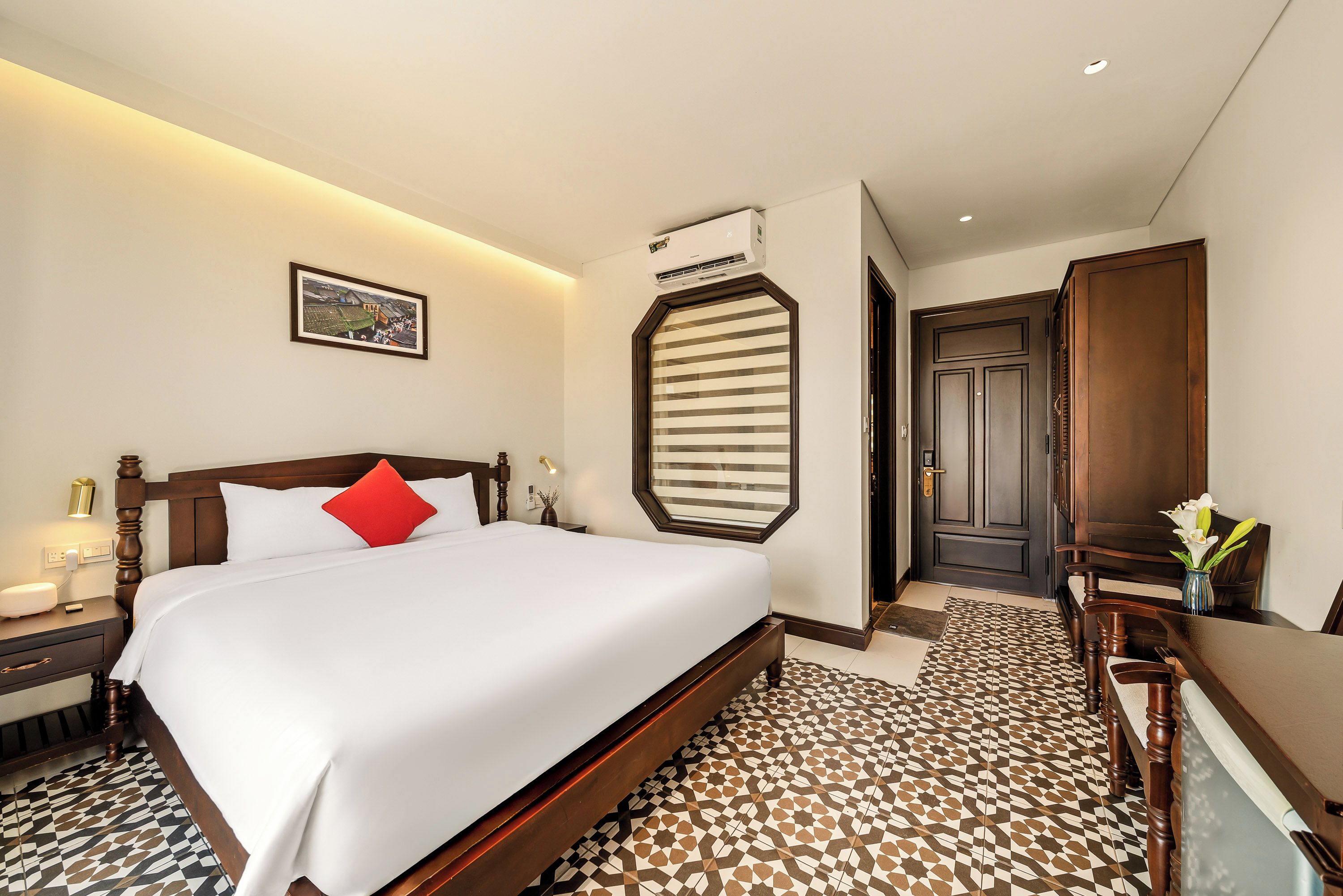 A hotel room with a large bed, a nightstand, a bedside table, a framed picture on the wall, a window with blinds, and a door leading to another room.