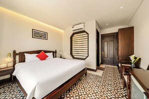 A hotel room with a large bed, a nightstand, a bedside table, a framed picture on the wall, a window with blinds, and a door leading to another room. - Manterra Hoi An Hotel (Da Nang)