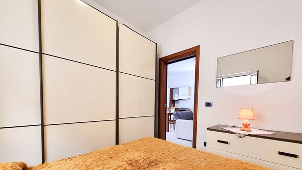 Ein Schlafzimmer mit Bett, Nachttisch mit Lampe, Spiegel und Blick zur Küche.