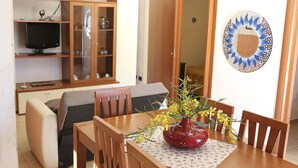 Ein Essbereich mit Tisch, Stühlen, einer Blumenvase, einem Fernseher auf einem Regal und einem Wanddekor.